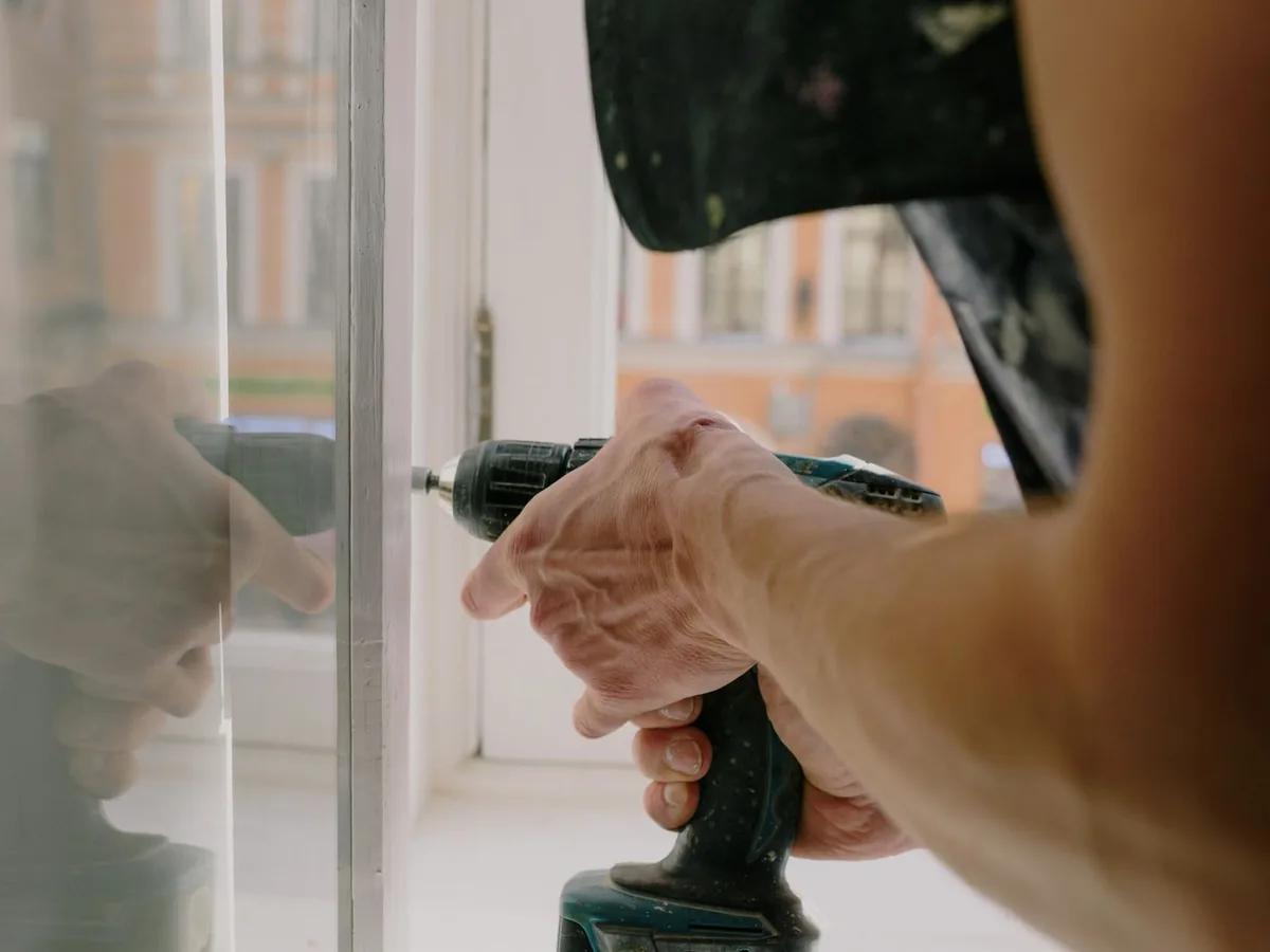 Hand driving a cordless drill into a white window frame